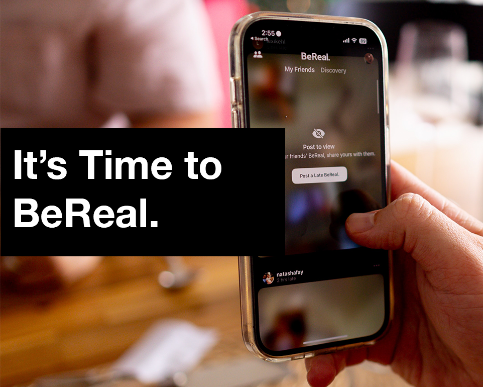 A person holds a phone with the BeReal app on the screen. Text in a balck box saying "It's time to BeReal"