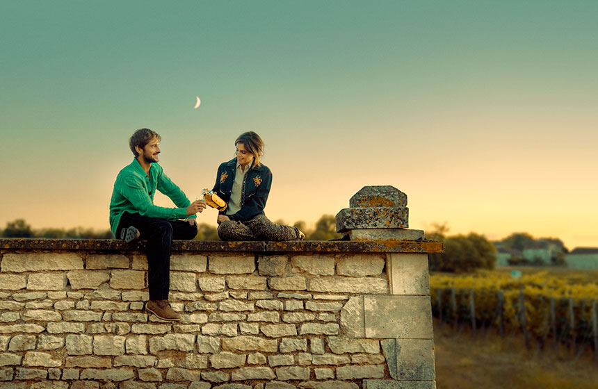 Two people sitting on a stone wall at sunset, holding a bottle.