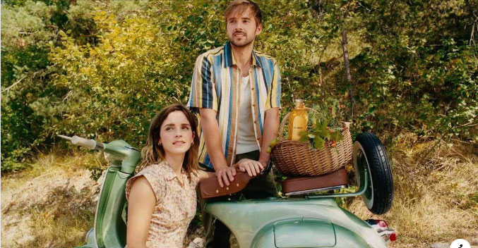 Two people sit on a vintage scooter with a picnic basket in a wooded area.